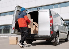 Man in colorful jacket loading cardboard boxes into a van outside an office building.